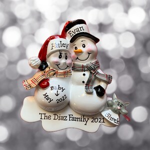 May include: A white snowman couple Christmas ornament with red, black, and white plaid scarves. The female snowman is wearing a red Santa hat and has the name "Haley" written on it. The male snowman has the name "Evan" written on his black hat. The snowman couple is holding a sign that says "Baby" with an arrow pointing down and "May 2022" written below. The ornament also has a gray cat with the name "Shrek" written on it. The base of the ornament says "The Diaz Family 2021".