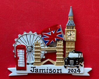 May include: A white and red Christmas ornament featuring iconic London landmarks, including Big Ben, the London Eye, a red telephone booth, and a black taxi. The ornament has a white banner with the text "Jamison 2024".