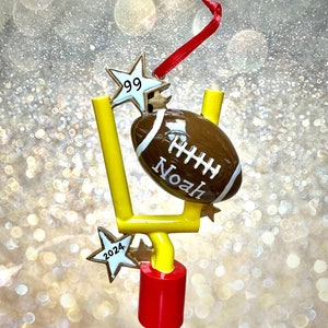 May include: A gold football goal post ornament with a brown football and two gold stars. The football has the name "Noah" and the number "99" on it. One star has the number "2024" on it.