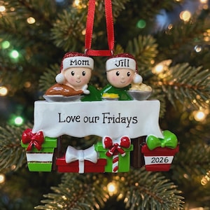 May include: A Christmas ornament featuring two figures in Santa hats labeled Mom and Jill sitting at a table with a white tablecloth printed with Love our Fridays. The base includes stacked gift boxes with red and green bows and the year 2026.