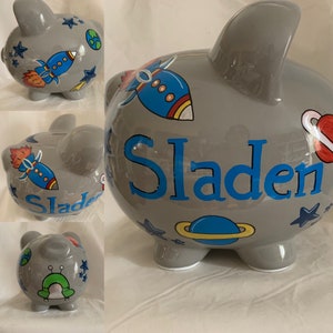 May include: Gray ceramic piggy bank with a space theme. The piggy bank is decorated with rockets, stars, planets, and the name "Sladen".