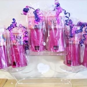 Puede incluir: Seis recipientes de plástico transparente llenos de caramelos coloridos, envueltos en cinta morada y blanca, y exhibidos en un soporte de acrílico transparente.