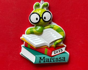 Op de afbeelding: Een groene en gele worm met een bril zit op een stapel boeken. De boeken zijn rood, geel en turquoise. Het onderste boek heeft de naam "Marissa" en het jaar "2025". De achtergrond is rood.