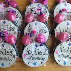 Puede incluir: Un conjunto de ocho maracas personalizadas con mangos rosas y ejes negros. Cada maraca está decorada con estrellas blancas, notas musicales y el nombre "Kylie's" con el texto "2nd Birthday".