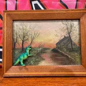 May include: A framed painting depicting a rural scene with a house, trees, and a path leading to a river. A green plastic dinosaur toy is positioned in the foreground.
