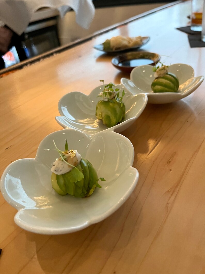 K&ouml;nnte beinhalten: Drei wei&szlig;e Keramikteller in Bl&uuml;tenform mit gr&uuml;ner Avocado und wei&szlig;er Sauce, die mit essbaren Goldflocken verziert ist. Die Teller stehen auf einer hellbraunen Holzoberfl&auml;che.