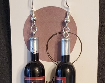 May include: A pair of handmade earrings featuring miniature wine bottle charms. The black bottles have silver tops and detailed labels with text and images. The earrings hang from silver-colored fish hook ear wires. The background includes a card that says "Handmade & Co."