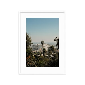 May include: A view of a city skyline with tall buildings and palm trees in the foreground. The sky is a light blue and the buildings are white.
