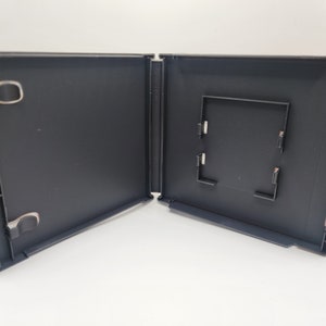 May include: An open, empty black plastic game case. The case is hinged and has a rectangular insert area. Metal clips are visible on the left side. The case is on a white surface.