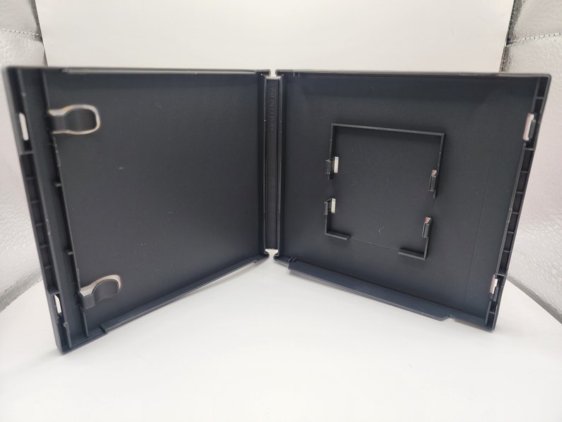 May include: An open, empty black plastic game case. The case is hinged and has a rectangular insert area. Metal clips are visible on the left side. The case is on a white surface.