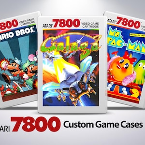 Könnte beinhalten: Drei Atari 7800 Videospielkassetten mit benutzerdefinierten Hüllen. Die Kassetten sind für die Spiele Mario Bros., Galaga und Ms. Pac-Man. Die Hüllen sind weiß mit einem schwarzen und roten Atari 7800 Logo.