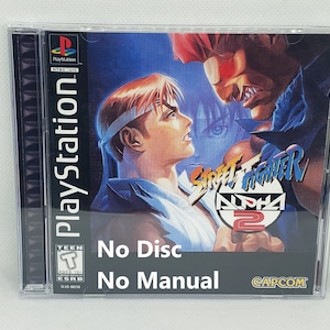 Może przedstawiać: Pudełko na grę PlayStation do Street Fighter Alpha 2. Pudełko jest wykonane z przezroczystego plastiku z czarnym grzbietem. Na przodzie pudełka znajduje się kolorowe zdjęcie dwóch postaci z gry. Tekst "Street Fighter Alpha 2" jest wydrukowany na czerwonym kółku białym kolorem. Tekst "Brak dysku" i "Brak instrukcji" jest wydrukowany czarnym kolorem u dołu pudełka.
