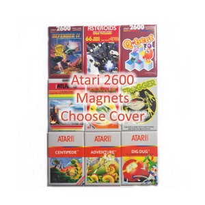 Puede incluir: Una colección de imanes de cartuchos de videojuegos Atari 2600. La imagen presenta varios títulos de juegos, incluyendo "Defender II", "Asteroids", "Q-bert", "Atlantis", "Frogger", "Centipede", "Adventure" y "Dig Dug". El texto "Atari 2600 Magnets Choose Cover" también es visible.