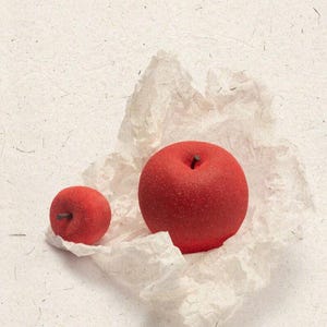 May include: Two red, apple-shaped objects rest on a crumpled piece of white paper against a textured, off-white background. The larger apple has a visible stem. The image includes Chinese characters and the text "HUI TIAU".