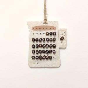 May include: A handmade calendar made of textured paper, featuring coffee bean date markers. The calendar displays the days of the week and numbered dates from 1 to 31. A small handle is on the side with the word "RESET". The calendar is suspended by a natural twine.