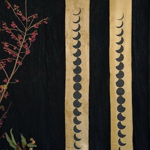 May include: Two vertical wall hangings with a beige background and a series of black crescent moon shapes. The hangings are suspended from wooden dowels. A black textured background and a branch with pink flowers are also visible.