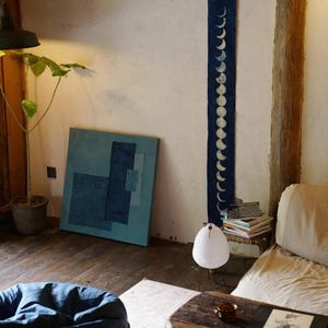 May include: An interior scene featuring a blue moon phase wall hanging, a blue abstract painting, and a white paper lamp. The room has wooden floors and a rustic wooden table. A beanbag chair is in the foreground.