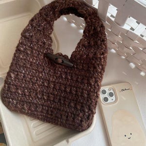 May include: A brown crocheted handbag with a wooden toggle closure. The bag is sitting on a beige container next to a phone case with a cartoon potato design. A white woven basket is in the background.