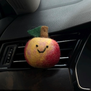 May include: A felt apple car air freshener with a smiling face. The apple is red and yellow with a green leaf and brown stem. The air freshener is placed on a car vent.