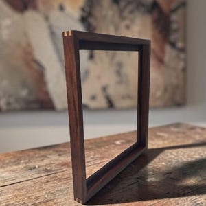 May include: A square wooden frame made of dark brown walnut wood with visible light-colored wood joinery at the corners. The frame has a deep profile and a smooth finish, showcasing the natural grain of the wood in a simple, open construction.