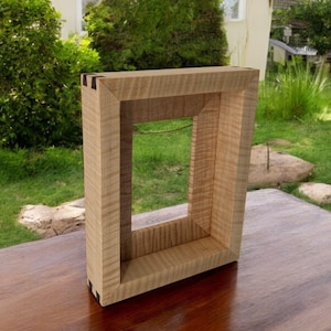 May include: A rectangular wooden picture frame crafted from light-colored tiger maple wood with a visible wavy grain pattern. The frame features dark, contrasting dovetail joinery at the corners, showcasing a clean, sturdy construction design.