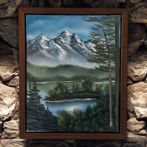 May include: A framed oil painting depicting a serene mountain lake scene. The painting features a small island with trees in the foreground, a lake with a reflection of the sky, and snow-capped mountains in the background.