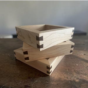May include: A stack of three square light-colored wooden boxes featuring visible dovetail joinery with dark brown accents at each corner. The boxes are constructed from smooth, natural-toned wood with a clear grain pattern and open tops.
