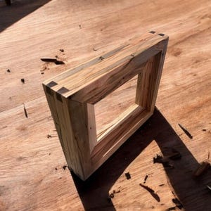 May include: A rectangular wooden picture frame crafted from light-colored wood with prominent dark grain patterns. The frame features visible dark wood dovetail joinery at the corners, creating a sturdy construction with a natural finish.