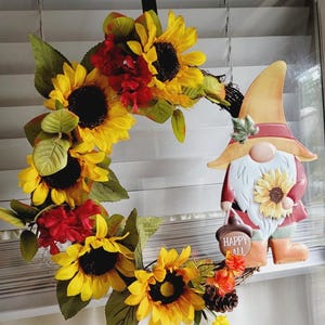 May include: A decorative wreath featuring bright yellow sunflowers, dark red floral accents, and green leaves. A gnome figure with a tall yellow hat, a white beard, and a brown acorn-shaped sign reading HAPPY FALL is attached to the right side.