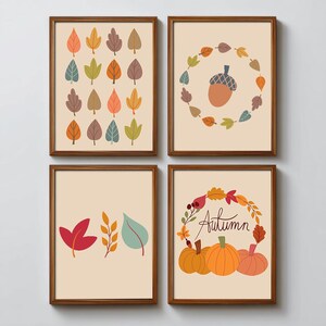 Op de afbeelding: Vier ingelijste kunstprints met herfstthema-illustraties. De prints tonen kleurrijke bladeren, een eikel omringd door bladeren, individuele bladeren en pompoenen met het woord "Autumn". De frames zijn bruin.