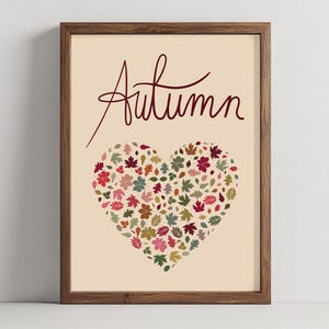 May include: A framed art print featuring the word "Autumn" in a cursive script above a heart shape. The heart is filled with colorful autumn leaves in shades of red, green, and brown, set against a beige background.