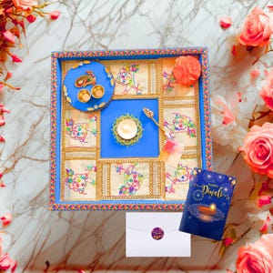 May include: A square blue decorative tray featuring embroidered fabric panels with colorful figures, a circular blue dish with gold accents, a central candle holder, a small spoon, and a greeting card that reads Happy Diwali.