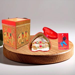 May include: A square wooden box with gold elephant motifs, a heart-shaped red container holding a gold bracelet with pink stones and pearls, and a red card featuring a peacock feather illustration and the text Happy Raksha Bandhan.