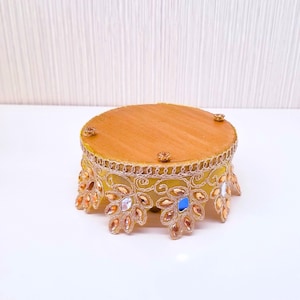 May include: A round, decorative pedestal in gold and yellow. The top is a solid gold color, while the sides are yellow with gold filigree and jewel accents. The pedestal has a scalloped edge with decorative jewels.