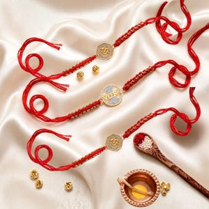 May include: Three red thread bracelets featuring gold-toned decorative centers. Two bracelets have octagonal charms with an Om symbol, and one features a circular charm with the name VEER in gold lettering. Small gold beads, a wooden spoon with rice, and a small diya lamp are included.