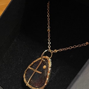 Peut inclure: Un collier dor&eacute; avec un pendentif en forme de goutte contenant un int&eacute;rieur sombre et paillet&eacute;. Le pendentif est entour&eacute; d'un cadre en m&eacute;tal dor&eacute;, incluant une fine barre transversale dor&eacute;e et un petit accent circulaire dor&eacute;, sur une cha&icirc;ne.