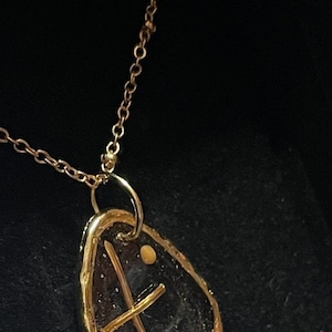 Peut inclure: Un collier pendentif dor&eacute; avec une pi&egrave;ce irr&eacute;guli&egrave;re en forme de goutte d'eau, en pierre ou en verre sombre, encadr&eacute;e de m&eacute;tal dor&eacute;. Une fine structure en croix dor&eacute;e recouvre la surface sombre, accompagn&eacute;e d'un petit accent dor&eacute; rond.