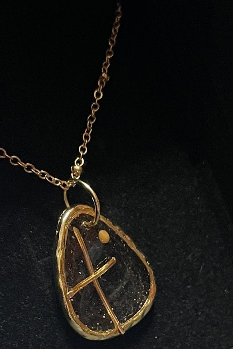 Peut inclure: Un collier pendentif dor&eacute; avec une pi&egrave;ce irr&eacute;guli&egrave;re en forme de goutte d'eau, en pierre ou en verre sombre, encadr&eacute;e de m&eacute;tal dor&eacute;. Une fine structure en croix dor&eacute;e recouvre la surface sombre, accompagn&eacute;e d'un petit accent dor&eacute; rond.