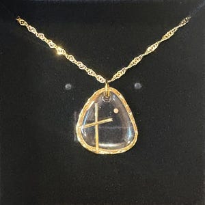 Peut inclure: Un collier dor&eacute; avec un pendentif transparent en forme de galet irr&eacute;gulier, incrust&eacute; d'une croix dor&eacute;e et d'un petit point. Le pendentif est entour&eacute; d'un bord dor&eacute; textur&eacute; et est suspendu &agrave; une cha&icirc;ne dor&eacute;e torsad&eacute;e.