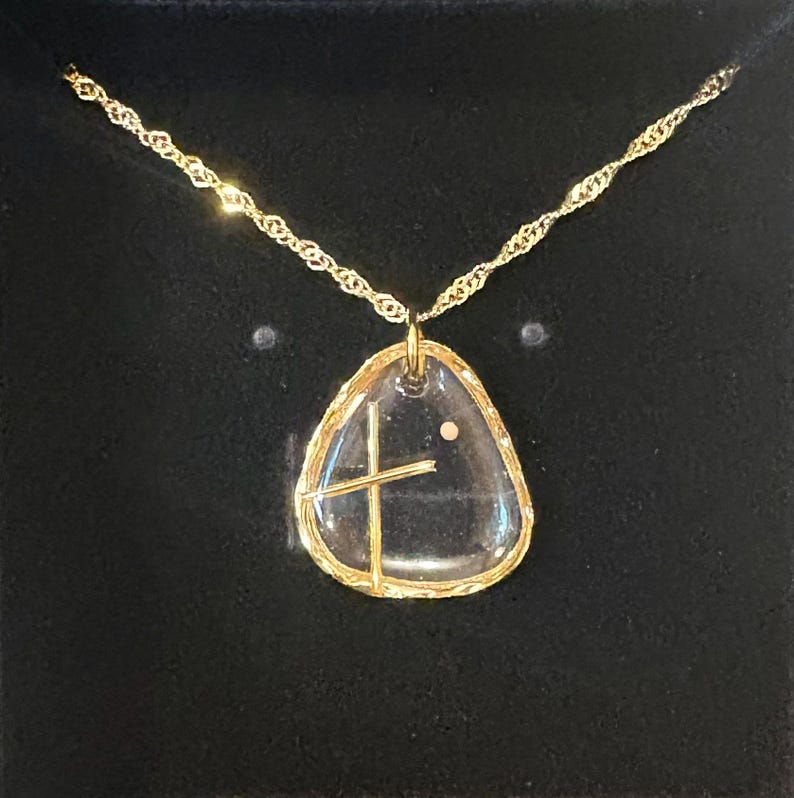 Peut inclure: Un collier dor&eacute; avec un pendentif transparent en forme de galet irr&eacute;gulier, incrust&eacute; d'une croix dor&eacute;e et d'un petit point. Le pendentif est entour&eacute; d'un bord dor&eacute; textur&eacute; et est suspendu &agrave; une cha&icirc;ne dor&eacute;e torsad&eacute;e.