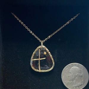 Peut inclure: Un collier dor&eacute; avec un pendentif au centre translucide, sombre et mouchet&eacute;, entour&eacute; d'un cadre dor&eacute; textur&eacute;. Une fine croix dor&eacute;e et un petit point dor&eacute; sont incrust&eacute;s dans le pendentif. Une pi&egrave;ce de 25 cents est visible pour l'&eacute;chelle.