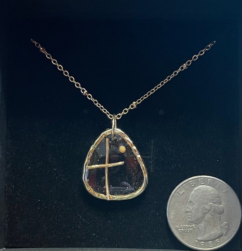 Peut inclure: Un collier dor&eacute; avec un pendentif au centre translucide, sombre et mouchet&eacute;, entour&eacute; d'un cadre dor&eacute; textur&eacute;. Une fine croix dor&eacute;e et un petit point dor&eacute; sont incrust&eacute;s dans le pendentif. Une pi&egrave;ce de 25 cents est visible pour l'&eacute;chelle.