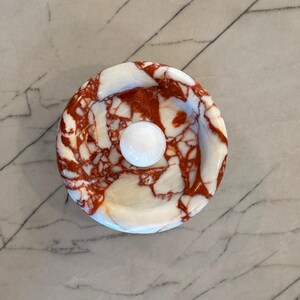 May include: A white marble dish with red and brown marbling. The dish is round and has a smooth surface. A white marble ball sits in the centre of the dish.