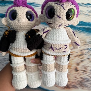 May include: Two handmade crochet dolls with purple hair buns and large, glittery eyes. One doll wears a black and white outfit with gold accents, the other a white top and gold chain. The dolls are set against a blurred ocean backdrop.