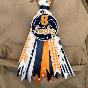 May include: A blue and orange volleyball charm with the number 8 and the name Ainsley on it. The charm is attached to a backpack with a variety of ribbons in orange, white, and blue.