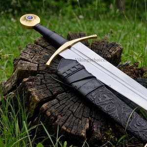 May include: A steel sword with a black grip, a gold-coloured crossguard, and a circular pommel featuring a blue stone inlay. The sword is accompanied by a black leather scabbard embossed with intricate scrollwork and gold-coloured metal accents.