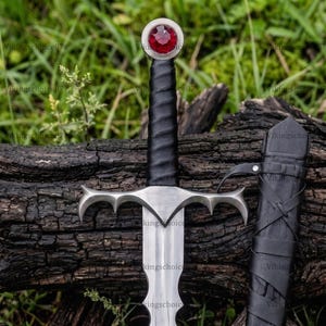 May include: A steel sword featuring a black leather-wrapped grip, a silver crossguard with pointed ends, and a circular pommel set with a faceted red gemstone. A matching black leather scabbard with crisscross lacing is positioned beside the sword.