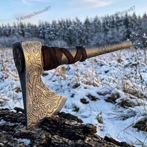 May include: A silver axe with intricate carvings and a brown leather-wrapped handle. The axe head features detailed patterns and a circular design. The axe is resting on a piece of wood in a snowy outdoor setting.