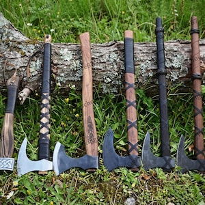 May include: A collection of seven Viking axes with wooden handles and metal blades. The axes vary in size and design, with some featuring intricate carvings and leather wrappings. The axes are displayed on a log in a grassy outdoor setting.