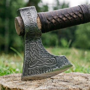 May include: A weathered metal axe with a wooden handle wrapped in dark brown leather. The axe head features engraved runic text and a Celtic knot design. The axe is resting on a tree stump.